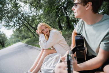 blurred man playing guitar while sitting on road near blonde girlfriend