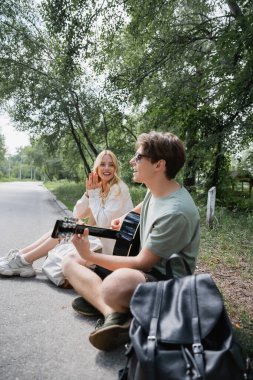 happy blonde woman applauding to boyfriend sitting on road and playing acoustic guitar