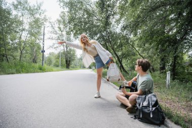 blonde woman stopping car near man sitting on road and playing guitar