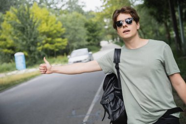 young hitchhiker in sunglasses stopping car on countryside road