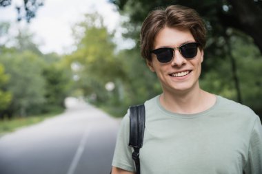 smiling tourist in sunglasses looking at camera on blurred road