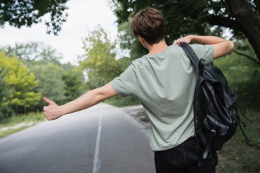 back view of hitchhiker with backpack stopping car with thumb up on blurred road