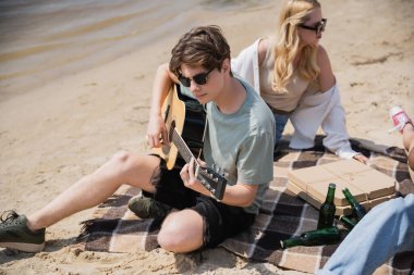man in sunglasses playing acoustic guitar during beach party with friends
