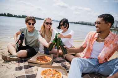 happy interracial friends in sunglasses clinking beer bottles during beach party