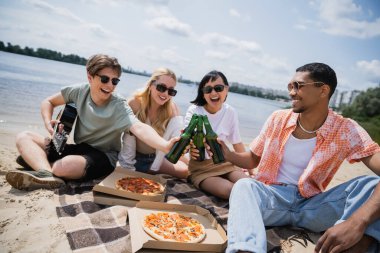 happy multiethnic friends in sunglasses toasting with beer near pizza on blanket
