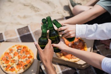 partial view of multiethnic friends clinking beer bottles near tasty pizza on beach