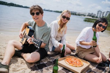 happy man playing guitar near happy interracial women during beach party