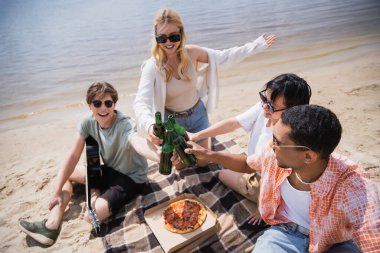 smiling woman pointing with hand while clinking beer bottles with multicultural friends