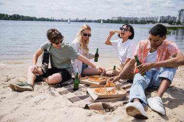 cheerful multicultural friends drinking beer and eating pizza on riverside