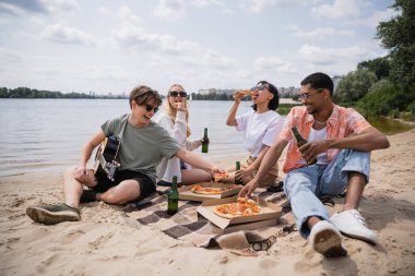 happy multicultural friends having beach picnic with beer and pizza