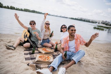 cheerful multiethnic friends waving hands during picnic on riverside
