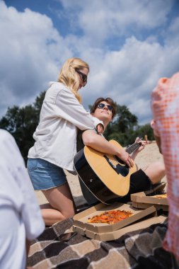 man playing guitar near blonde woman and friends during beach party
