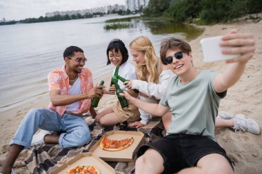 smiling man taking selfie with multiethnic friends clinking beer bottles near pizza