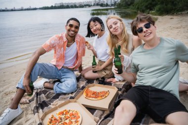 young multiethnic friends looking at camera during beach party with beer and pizza