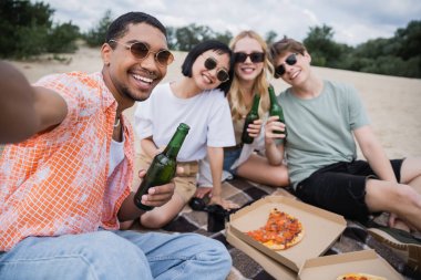 happy multiethnic friends in sunglasses holding beer near tasty pizza on beach