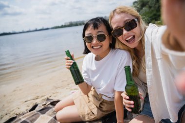 excited multiethnic women in sunglasses holding beer and looking at camera on beach