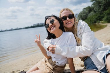cheerful asian woman showing victory gesture near blonde friend on beach