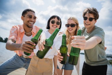 excited multicultural friends in sunglasses toasting with fresh beer on beach