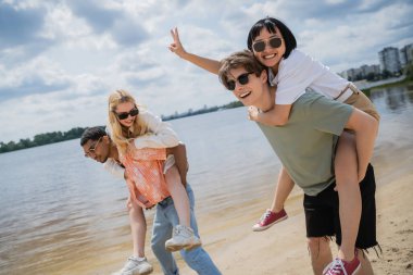 happy asian woman showing victory sign while having fun with interracial friends on beach