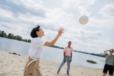 excited asian woman playing volleyball with interracial men on sand beach