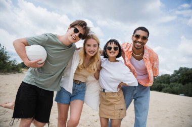 cheerful multiethnic friends in sunglasses smiling at camera on beach