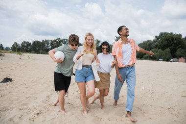 excited multicultural friends walking on sand beach with ball clipart