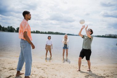 young man passing ball while playing beach volleyball with interracial friends clipart