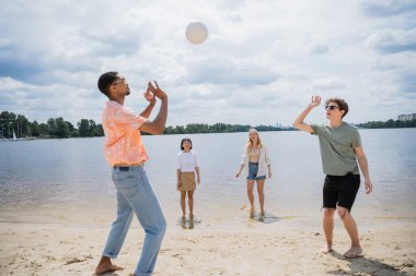 young multicultural friends playing beach volleyball near river