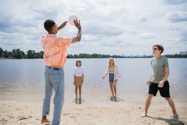 african american man passing ball while playing beach volleyball with multiethnic friends