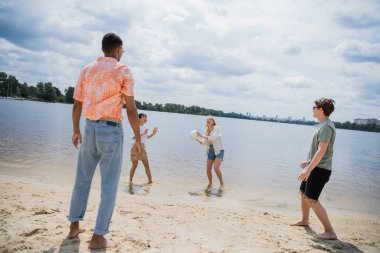 cheerful woman with ball standing in water while playing with interracial friends