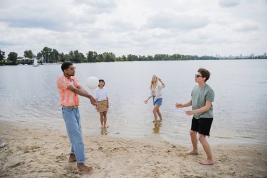 interracial men playing beach volleyball near women standing in water