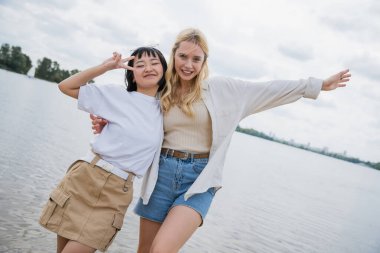 happy blonde woman embracing asian friend showing victory sign while posing near river