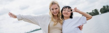 happy asian woman showing peace sign near blonde friend, banner