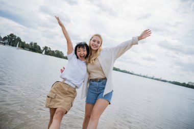 excited interracial friends hugging and posing with raised hands near river
