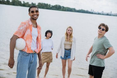 african american man with ball smiling at camera near multiethnic friends on beach