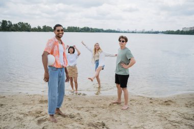 african american man holding ball and smiling at camera near interracial friends on beach