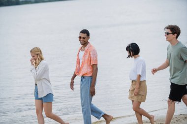 cheerful interracial friends walking along riverside