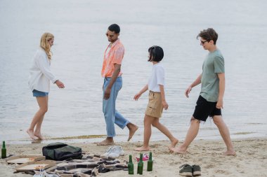 multicultural friends walking near beer bottles and pizza on beach
