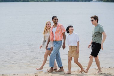 smiling multicultural friends looking at young man while walking along river