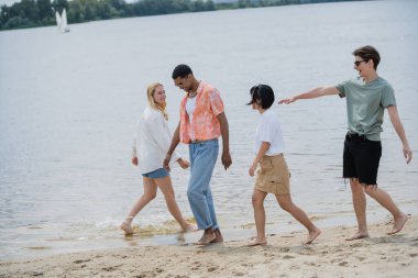 young man in sunglasses pointing with hand near multiethnic friends walking on beach