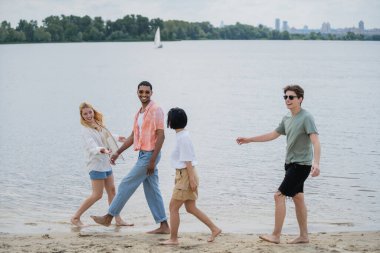 young barefoot multiethnic friends smiling while walking along river