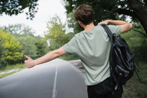 back view of hitchhiker with backpack stopping car with thumb up on blurred road