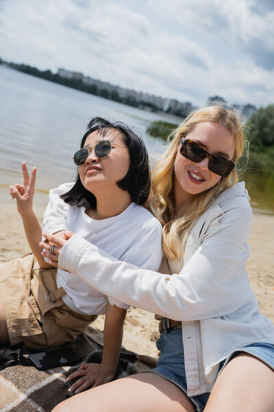 blonde woman in sunglasses embracing asian friend showing victory sign on beach