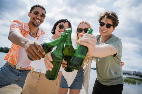 joyful multicultural friends toasting with beer bottles on riverside