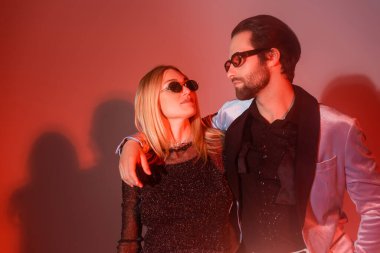 man in silk blazer and trendy eyeglasses embracing woman in black lurex dress on red background