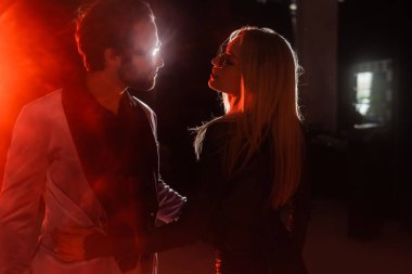stylish couple in eyeglasses looking at each other in darkness with bright light