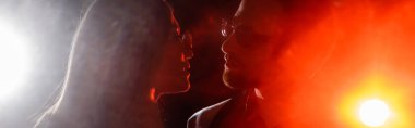 side view of couple in eyeglasses looking at each other on black background with bright light, banner