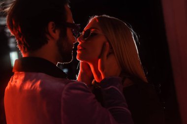 bearded man embracing neck of glamour woman in eyeglasses in darkness with light