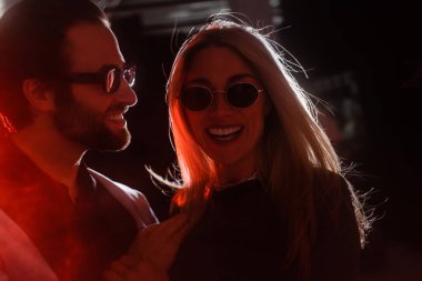 Smiling man in sunglasses hugging stylish girlfriend during party with lighting 