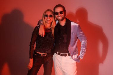 Stylish man hugging smiling girlfriend in sunglasses on red background